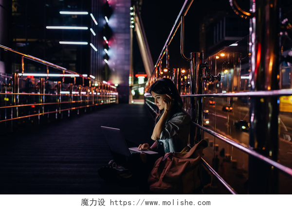 一个女人坐在黑夜的街边用着电脑晚上在城市街道上使用笔记本电脑时, 女性在耳机中听音乐的侧面视图